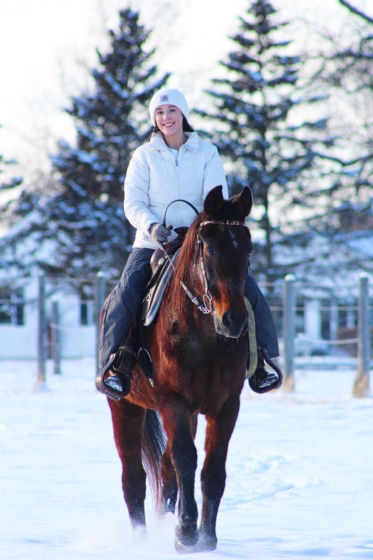 A rider on horseback trots through a snowy winter landscape with evergreen trees in the background.