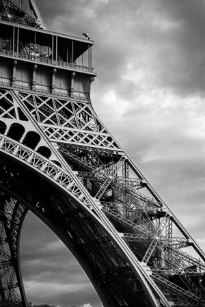 Architectural detail of the Eiffel Tower's iron lattice structure shown in dramatic black and white against cloudy sky.
