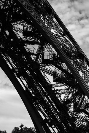 A dramatic black and white photograph showing the curved architectural details of the Eiffel Tower against a cloudy sky.