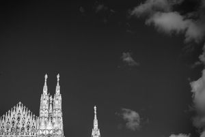 High contrast black and white photograph showing the ornate spires of Milan Cathedral against a dark sky with clouds.