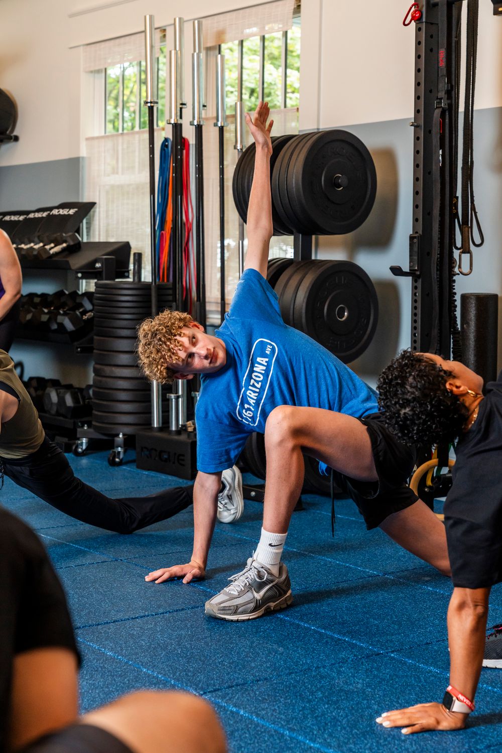 Progression of a person performing a hip mobility exercise on a blue gym floor next to weight racks.