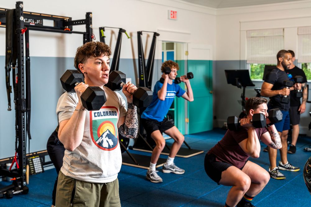 People work out with dumbbells and weights during a fitness class in a bright blue-walled gym space.