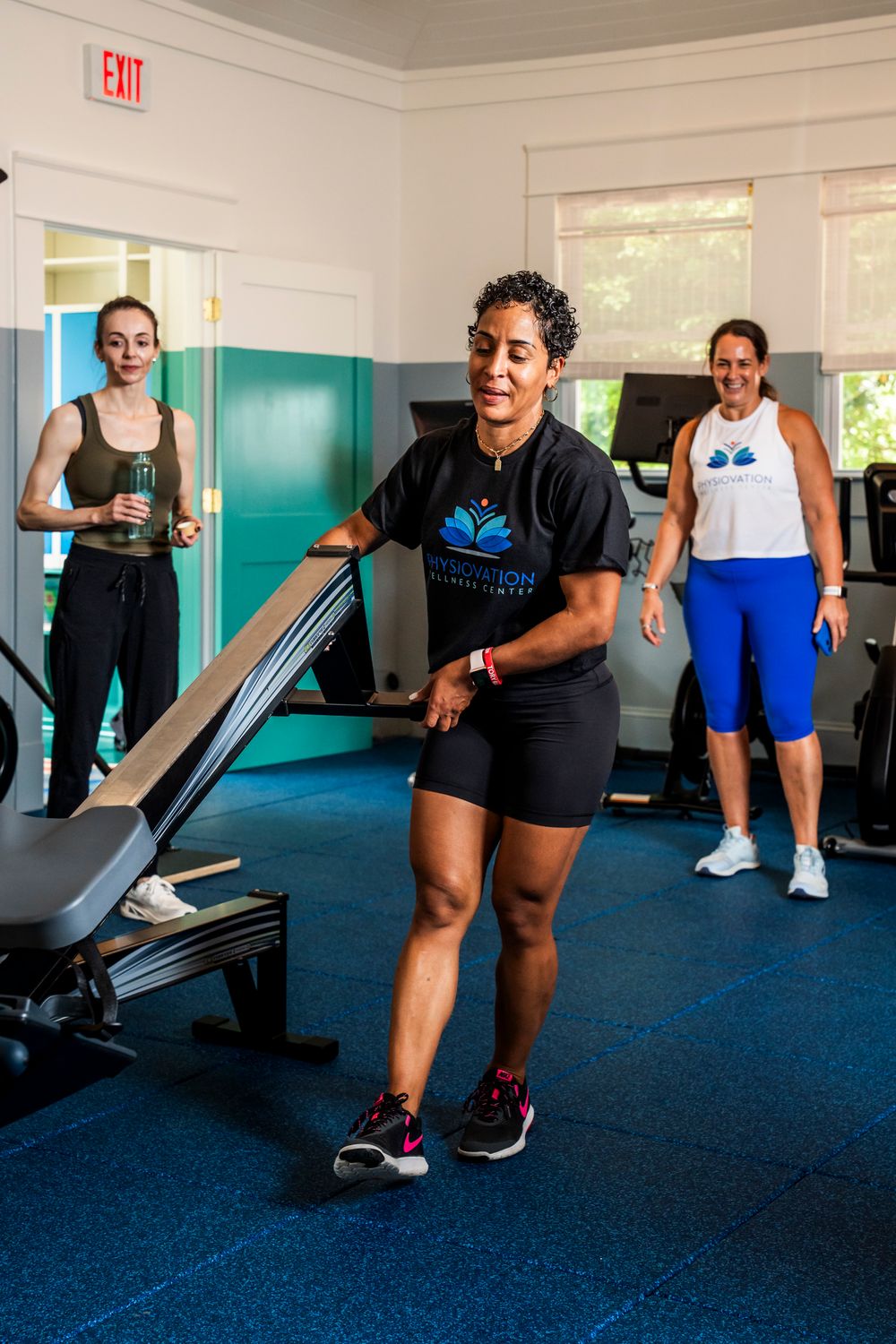 People exercising and doing strength training with equipment in a fitness room with blue carpet and turquoise walls.