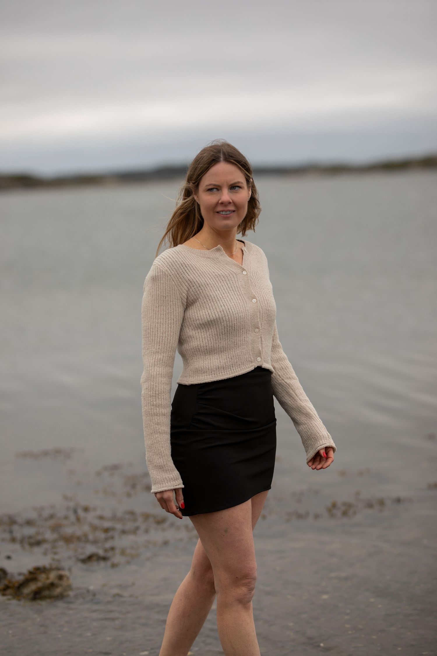 kvinna står i vattnet i strandkanten. hon har en svart klänning och en beige kofta. i bakgrunden syns havet.