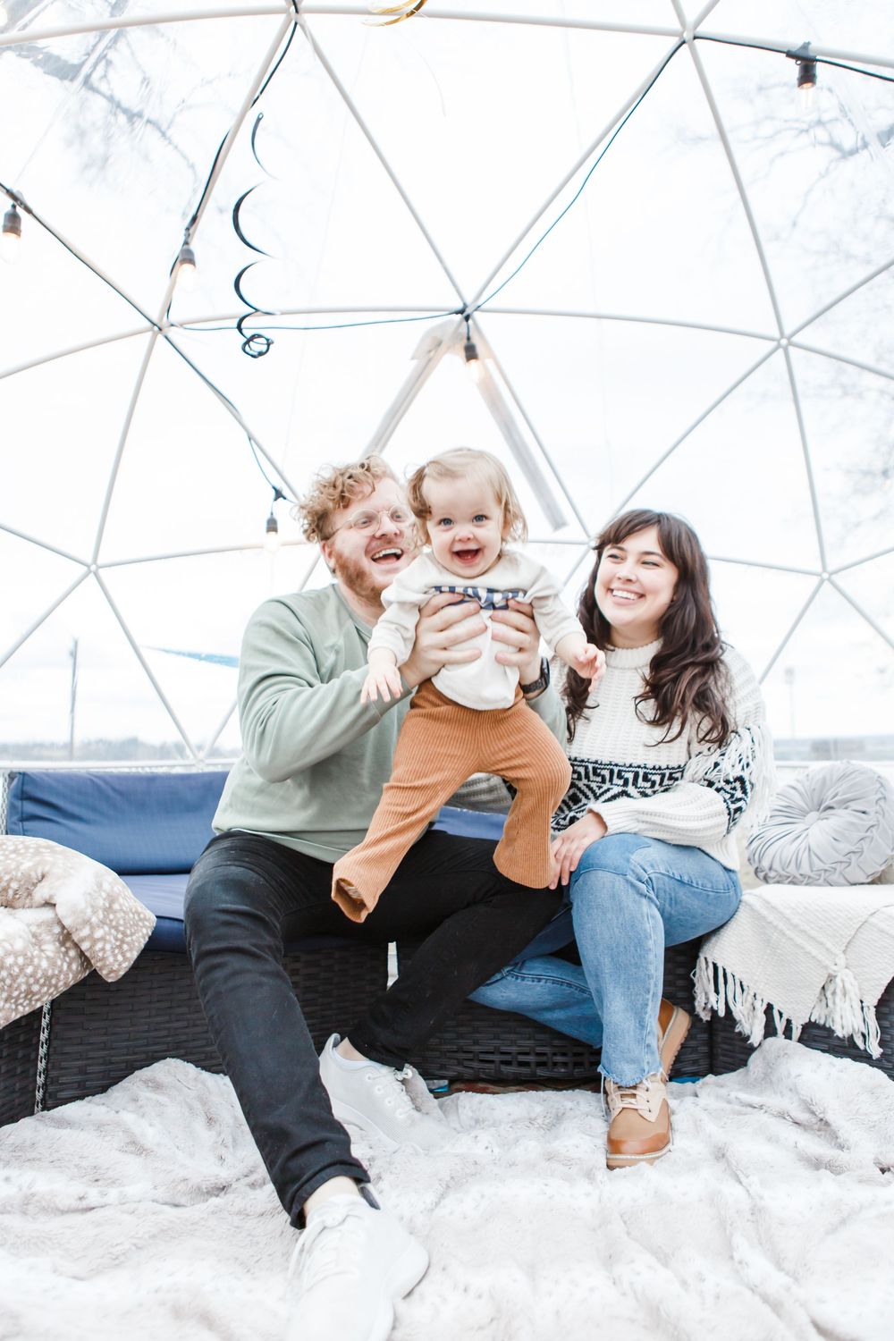 A small group enjoys a cozy moment together inside a geodesic dome with white blankets and cushions.