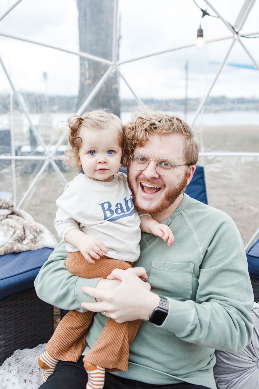A parent and child share joyful moments together while sitting outdoors with a bright white geometric structure behind them.