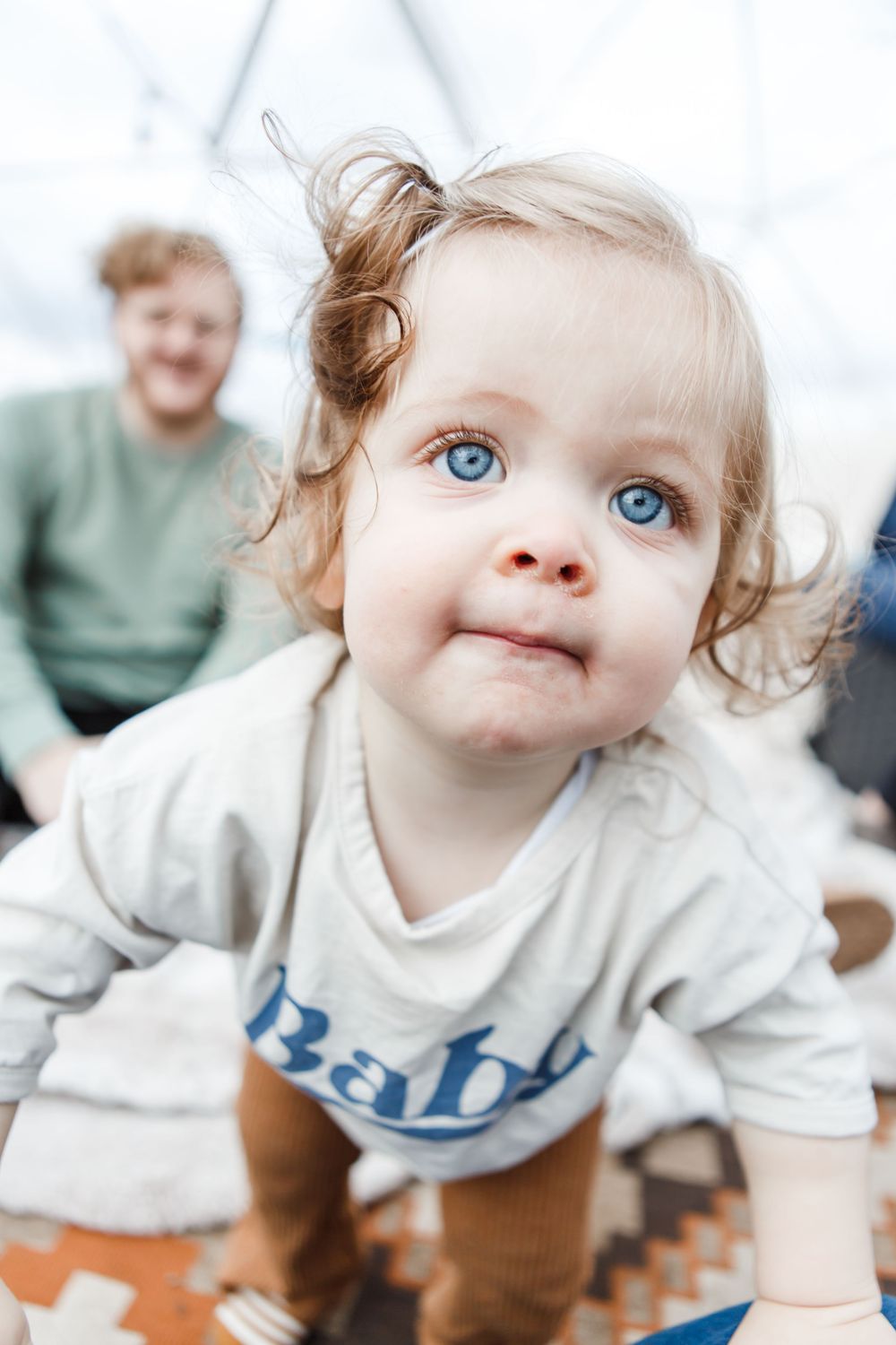 Young child in a white shirt and brown pants leans forward while looking up with bright blue eyes.