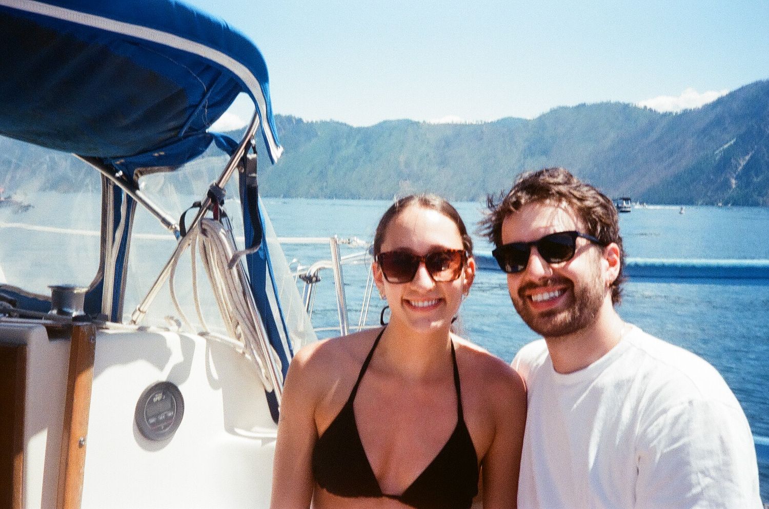 Couple enjoying a sunny day on a boat with mountain lake scenery in the background.