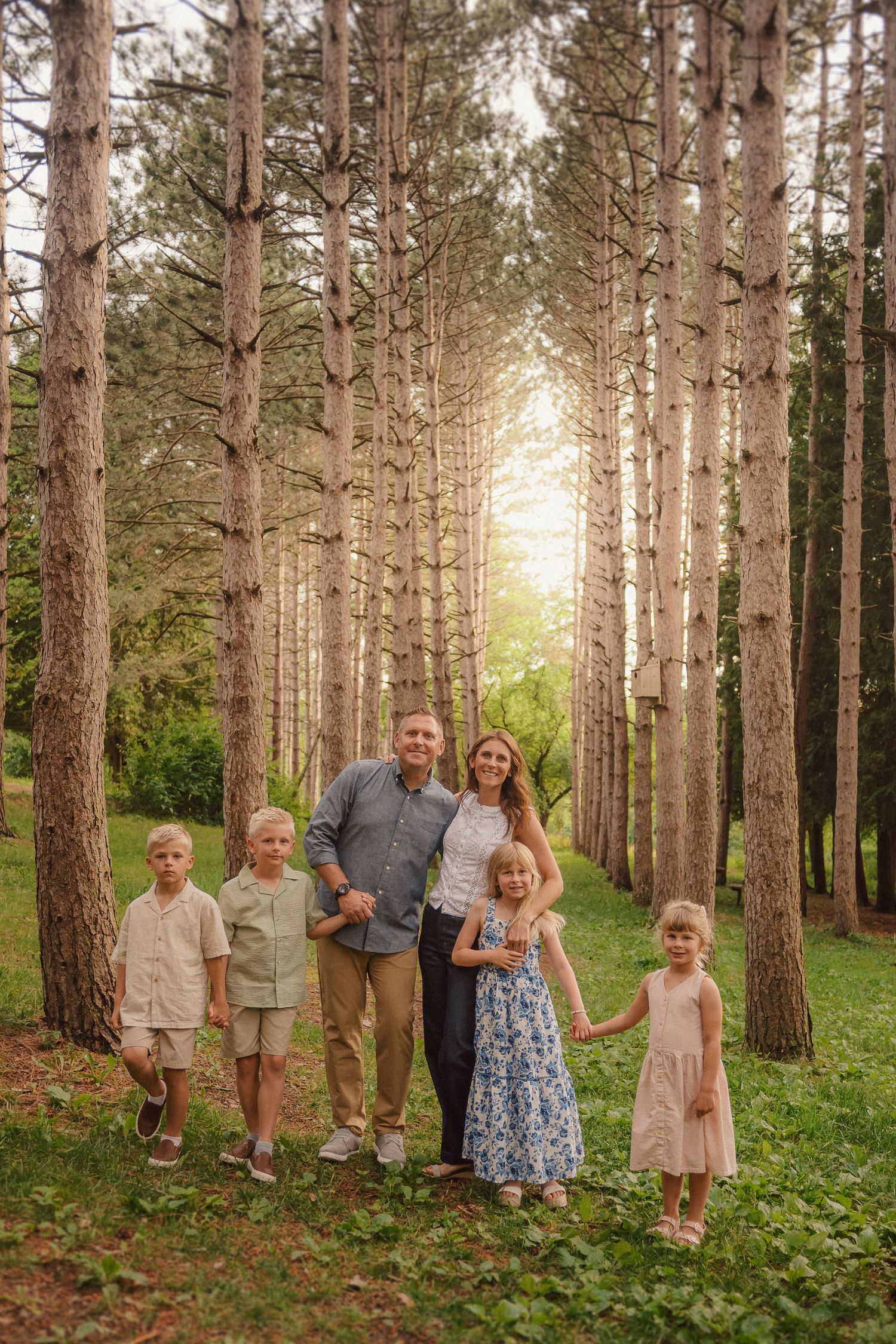 A family poses under towering pine trees in a forest during golden hour sunlight.