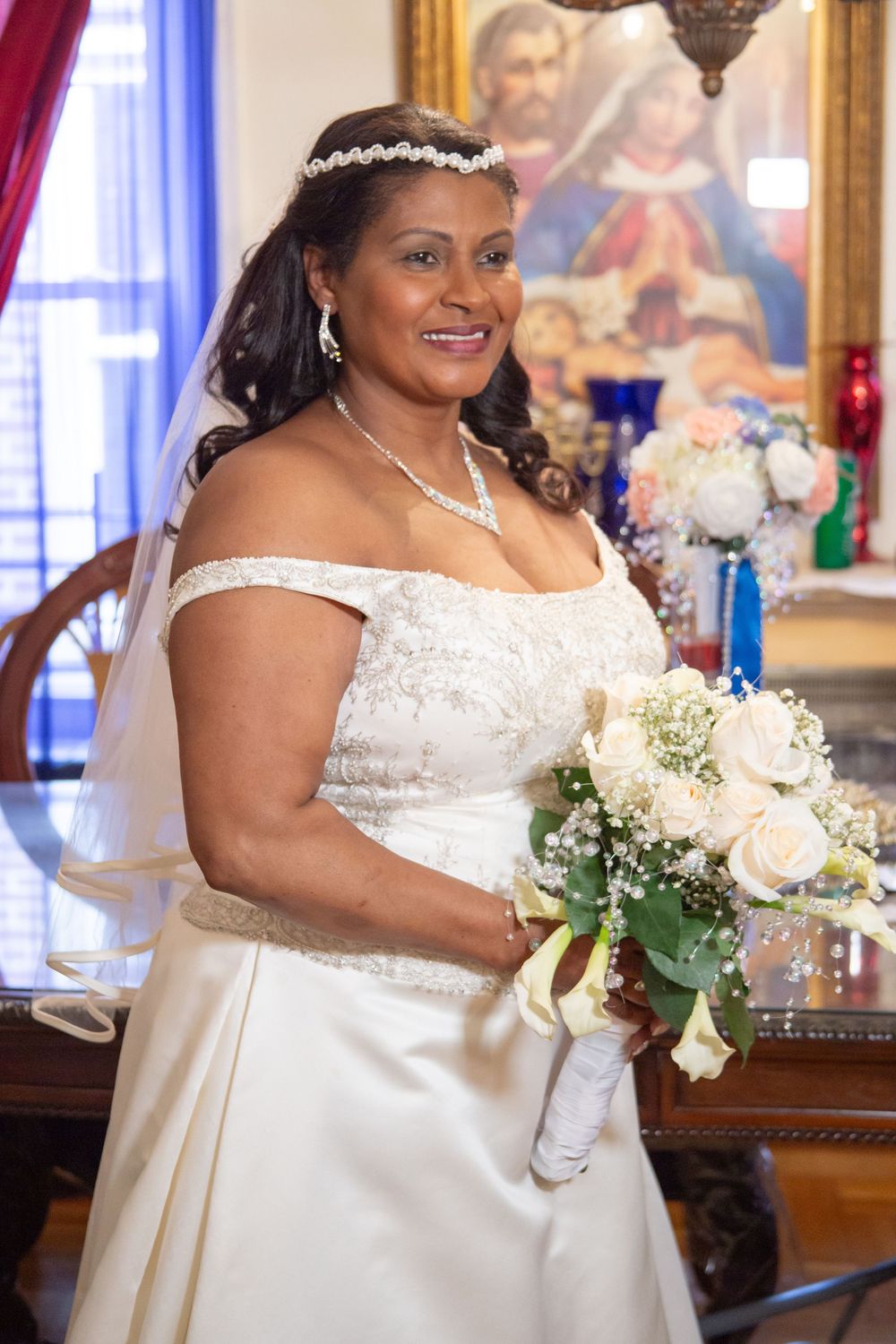 A bride wearing an elegant off-shoulder white satin wedding dress holds a cream and white bouquet with delicate greenery.