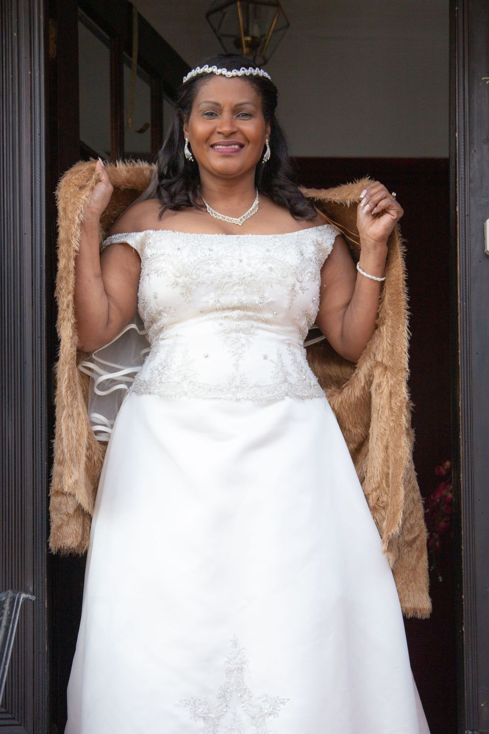 Bride wearing a white wedding dress with delicate details and tiara stands in doorway wrapped in gold shawl.