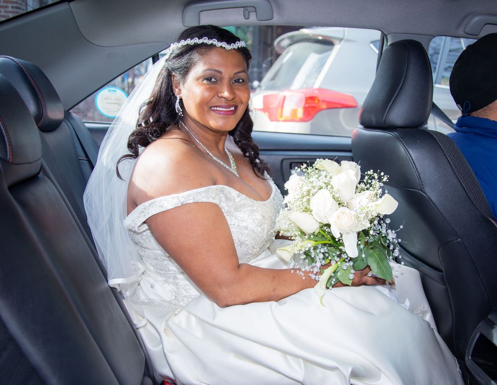 A person wearing a white wedding dress sits in the backseat of a car with a bouquet of white flowers.