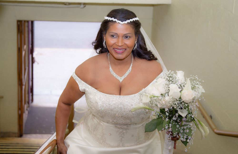 A bride in an off-the-shoulder white wedding gown smiles while holding a white bouquet.
