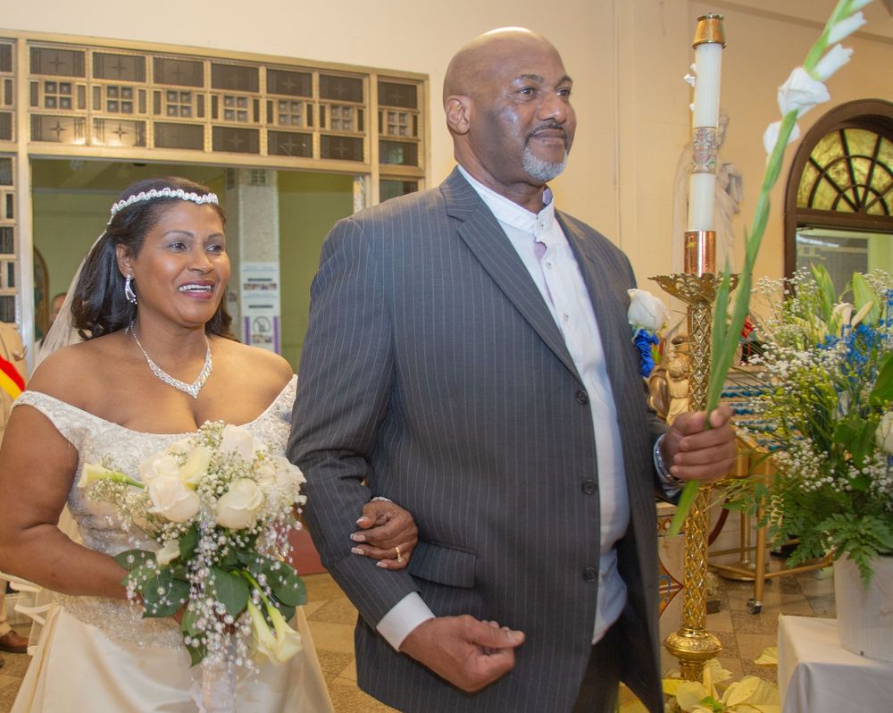 Two people in wedding attire smile while walking down the aisle together with floral arrangements visible.
