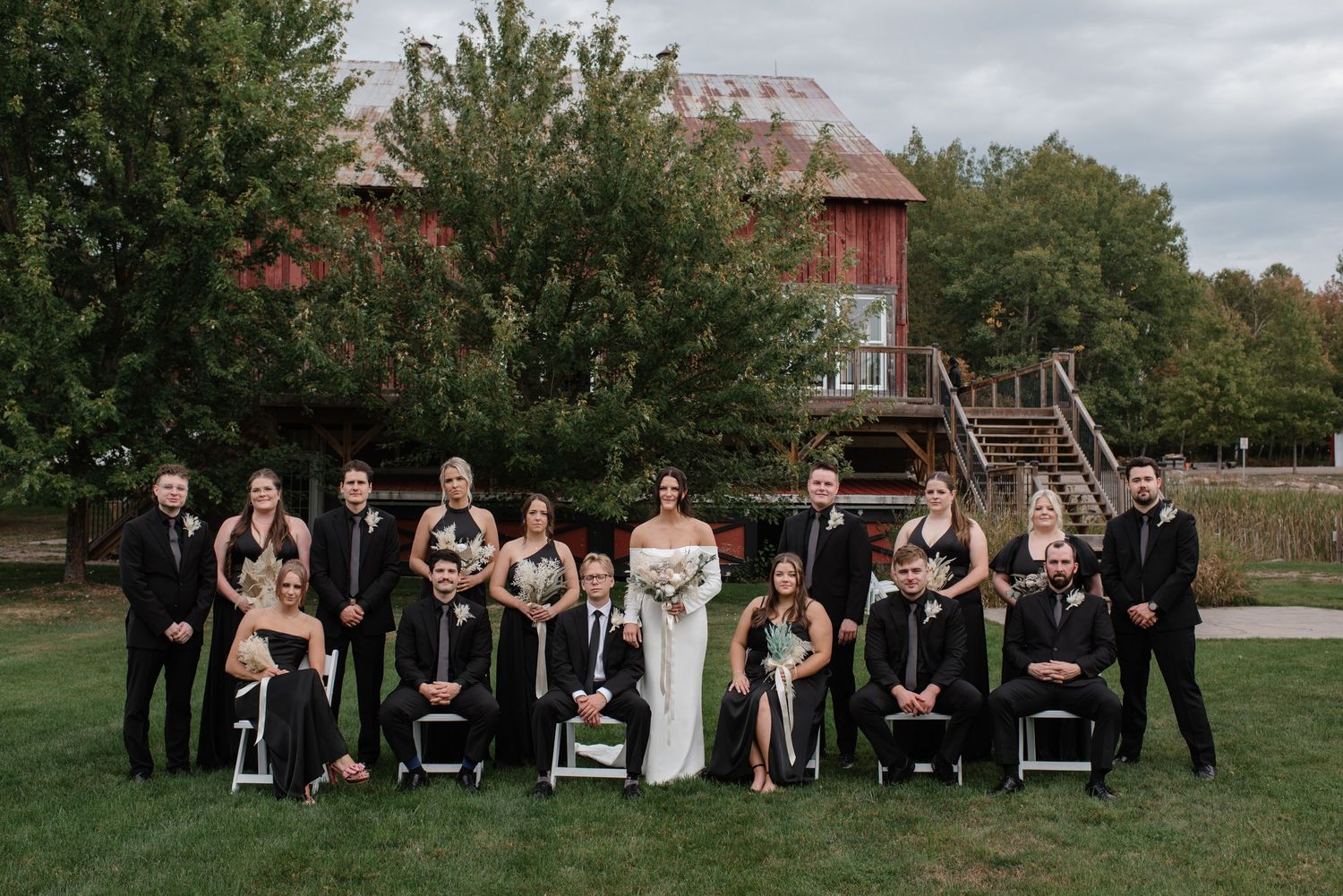 A wedding party group poses outdoors on a lawn with a rustic red barn and trees in the background.