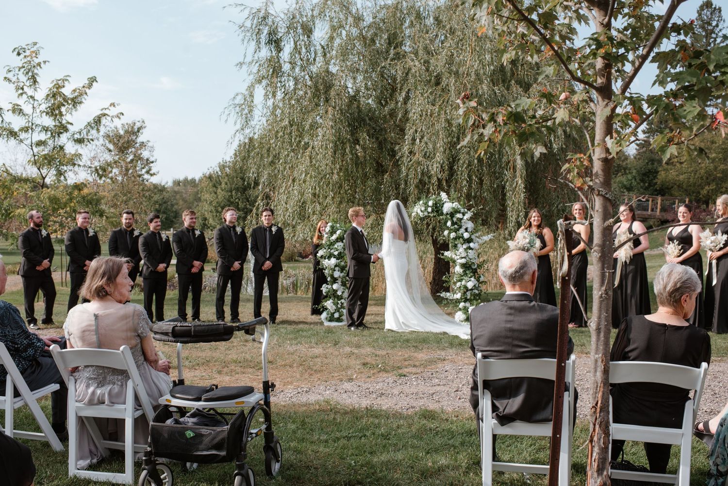 An outdoor wedding ceremony takes place under a weeping willow tree with white chairs and floral arrangements.