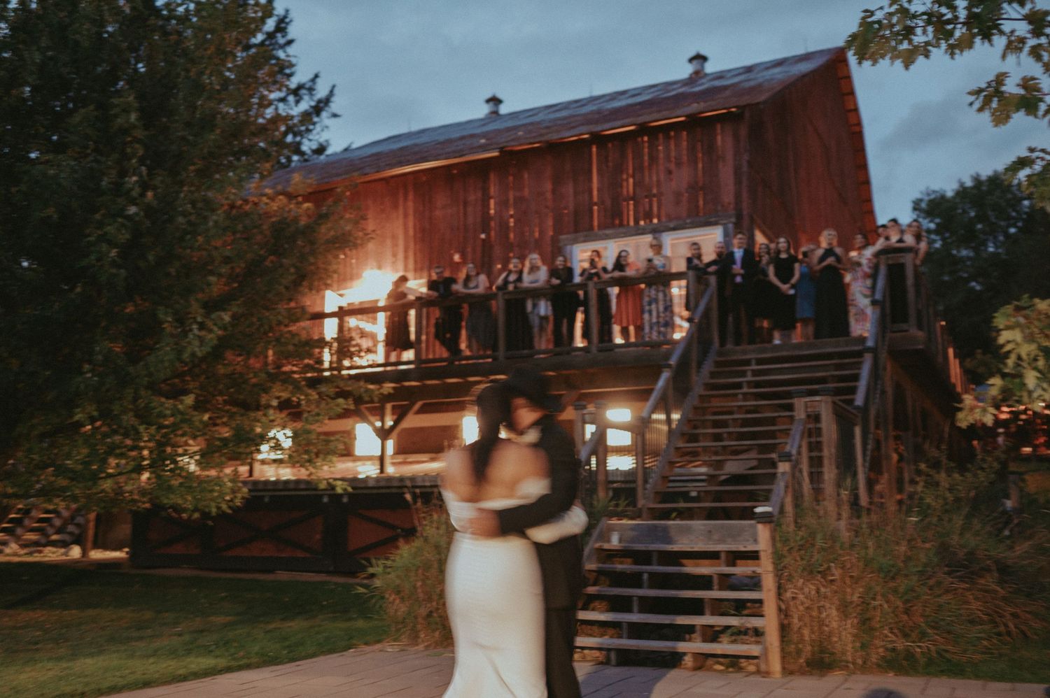 A couple shares a romantic moment outside a rustic barn venue while guests watch from the staircase at dusk.