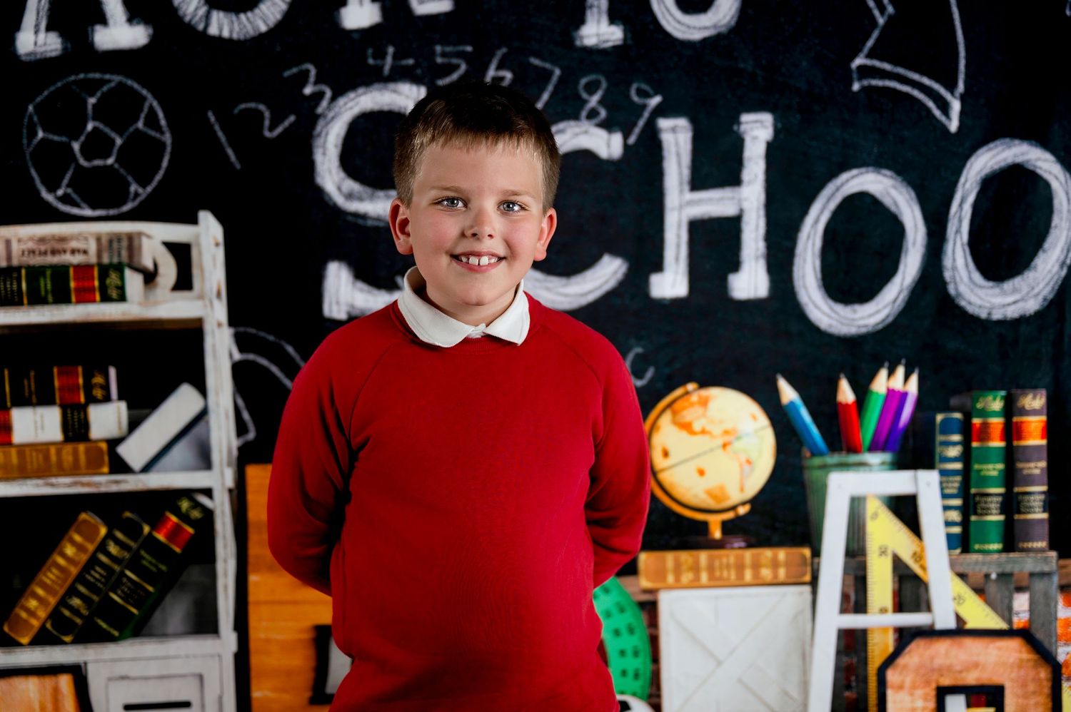 Student in red sweater stands in front of school chalkboard with supplies and globe in classroom setting.