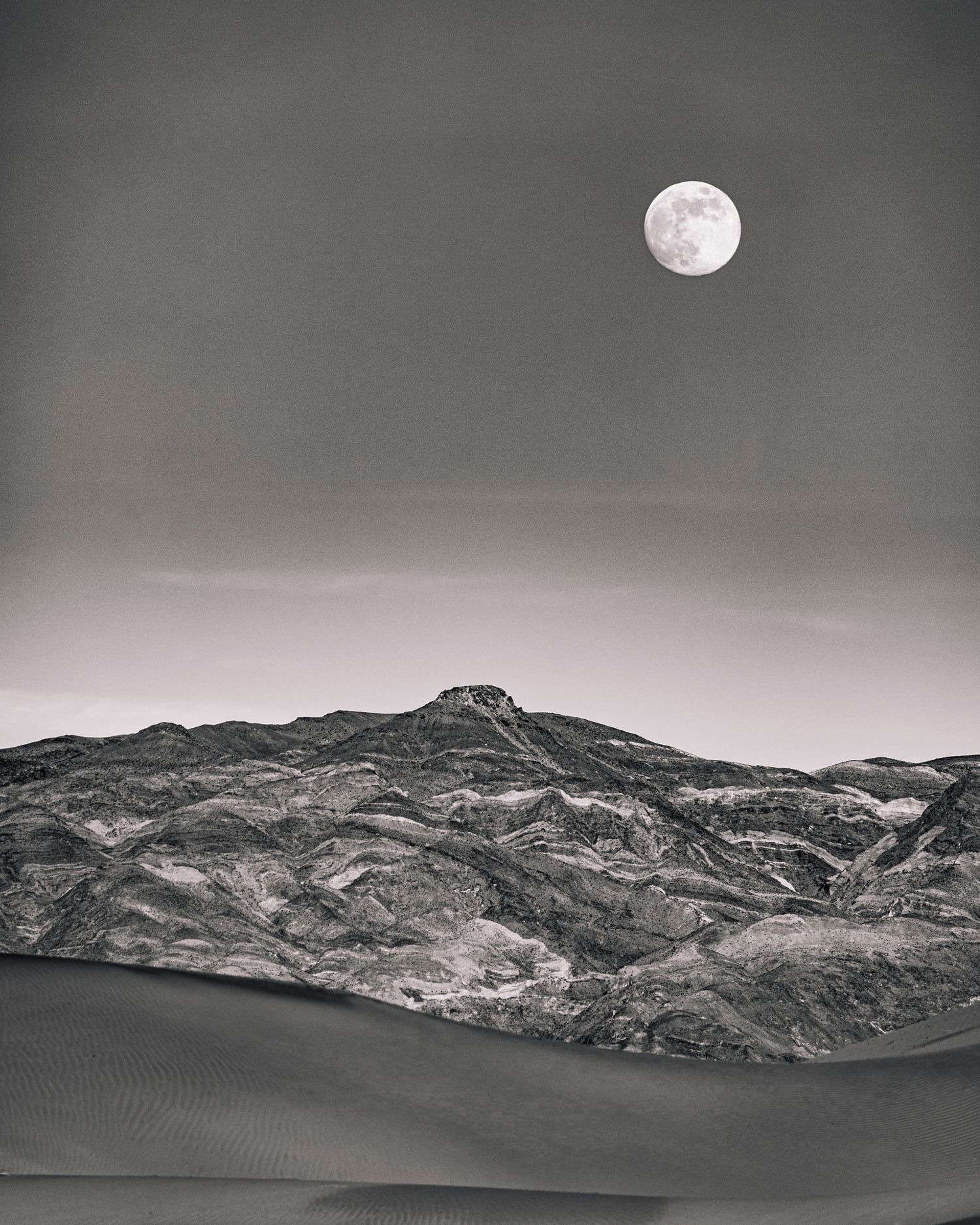 Full moon rising over dramatic desert mountain range above sand dunes in black and white.