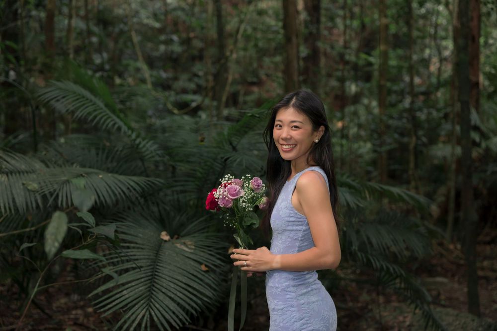 Woman in blue dress holding flowers while standing in a lush forest setting.