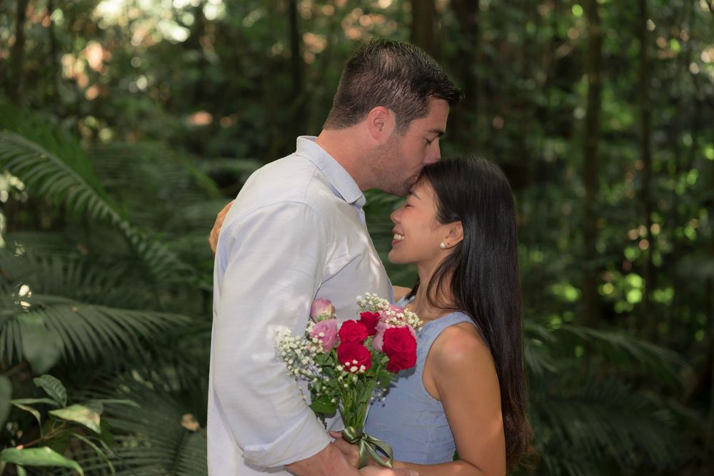 Couple sharing romantic moment while holding flowers in a tropical forest setting.