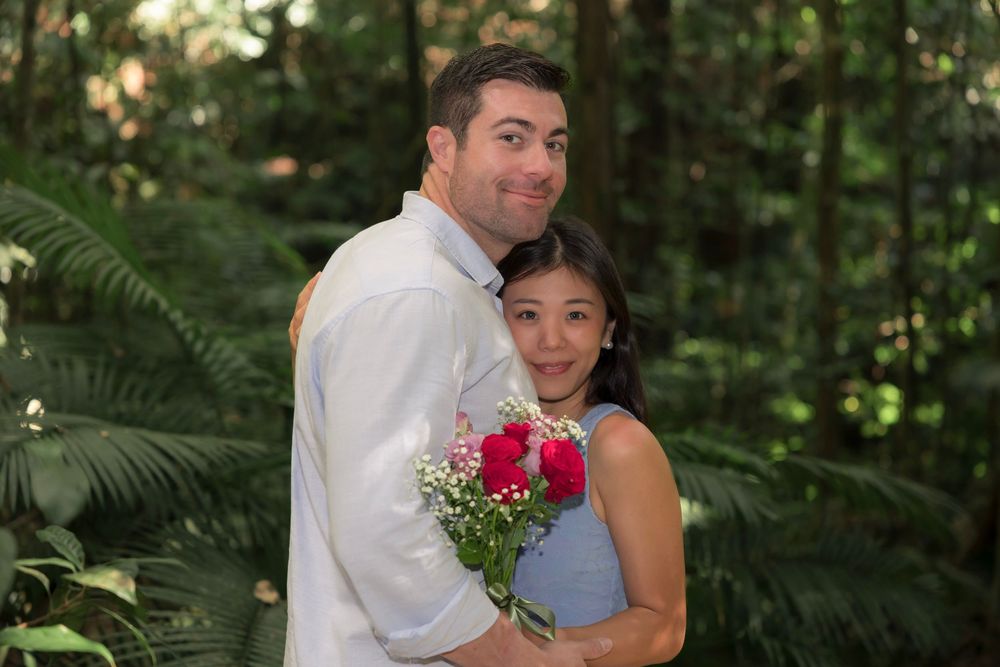A romantic couple poses in a lush garden setting with red roses and white baby's breath flowers.