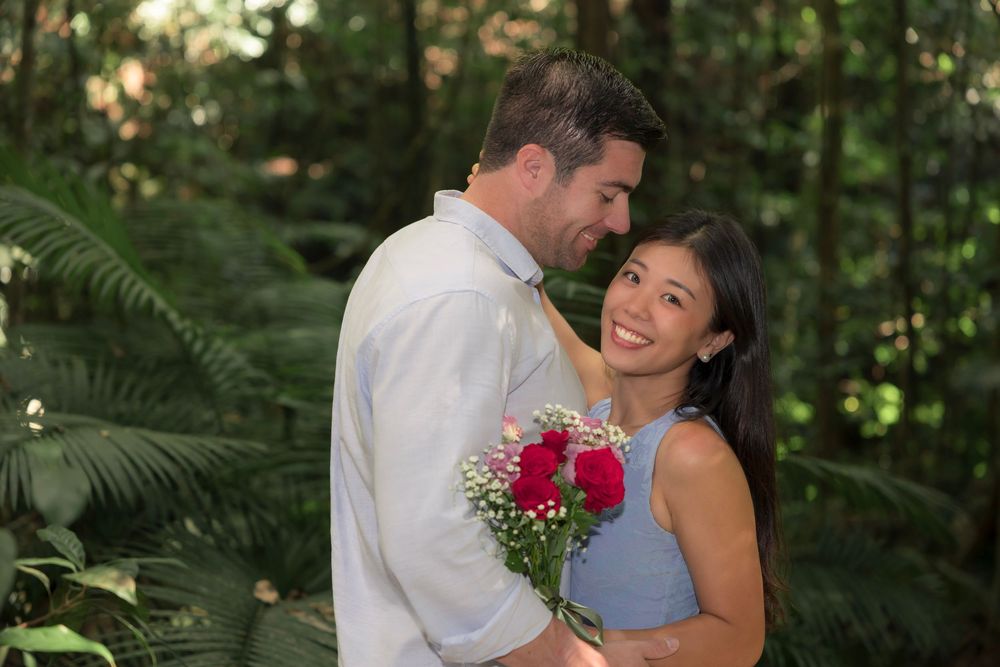 A romantic couple embraces in a lush green forest setting while holding red flowers.