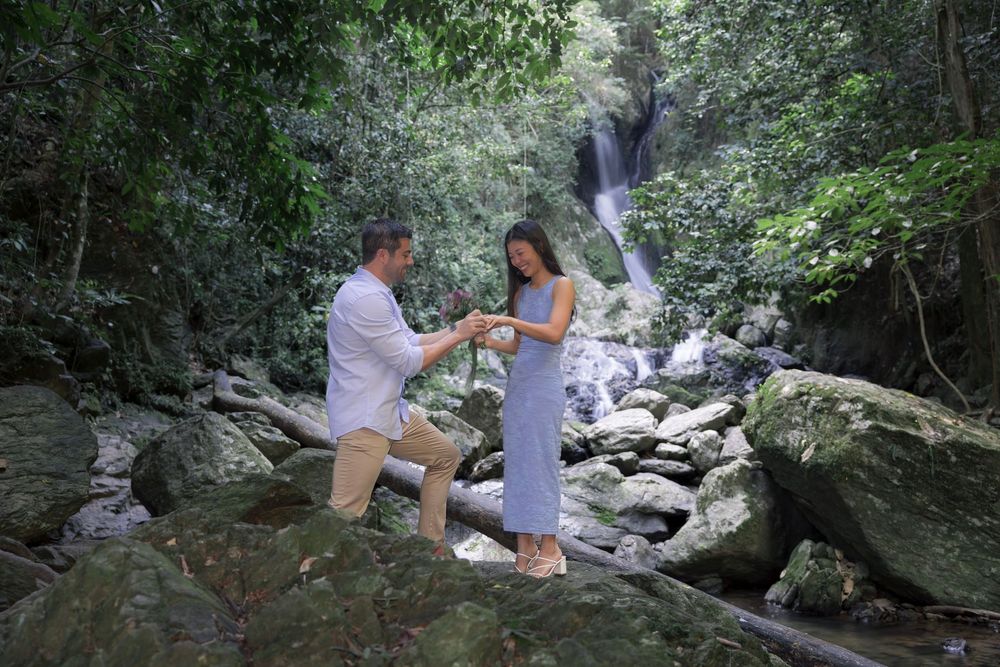 A couple sits beside a flowing stream in a peaceful forest setting, surrounded by large rocks and lush greenery.