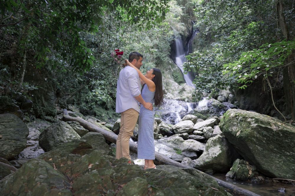 A couple stands on rocky terrain near a cascading waterfall in a lush forest setting.