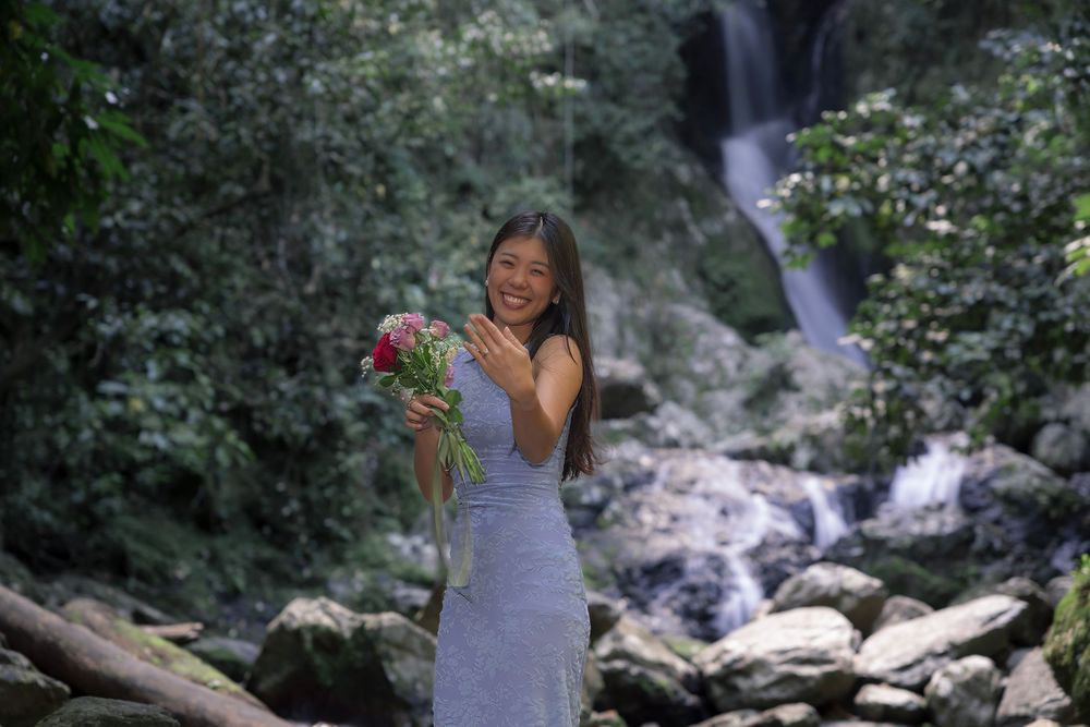 A person holds a vibrant bouquet in a light dress while standing near a cascading waterfall in a lush forest setting.