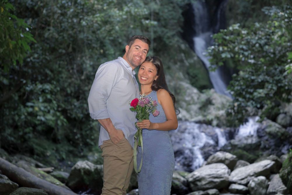 Romantic couple poses for waterfall engagement photos in lush green forest setting.