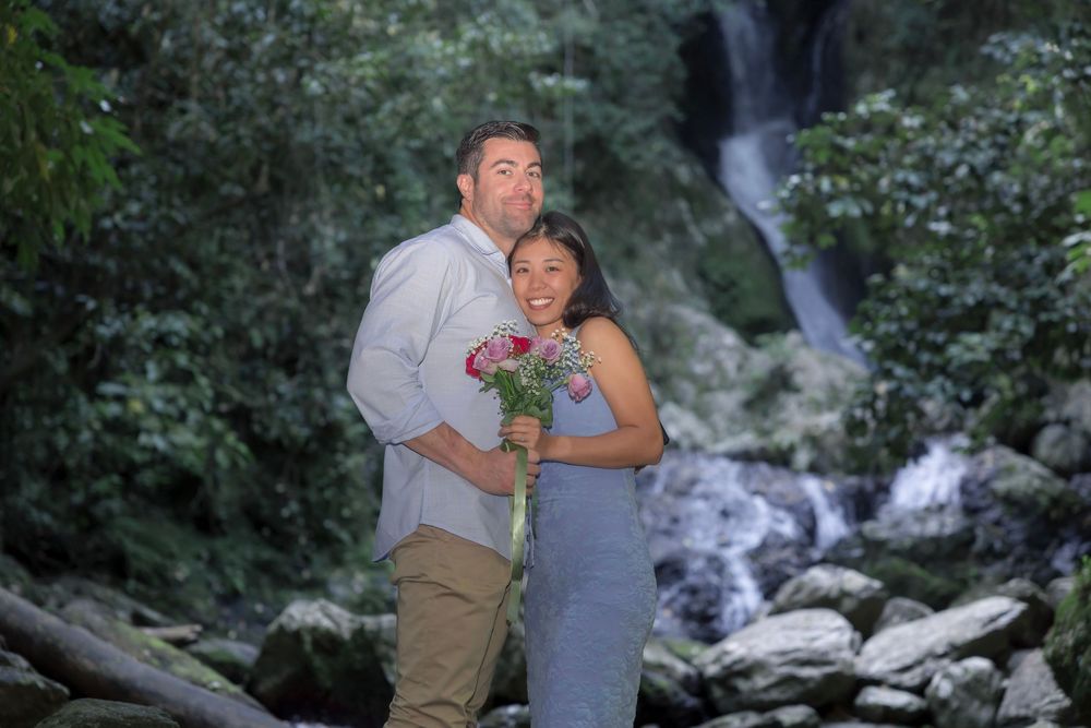 A couple poses together by a cascading waterfall surrounded by lush greenery and rocky terrain at sunset.