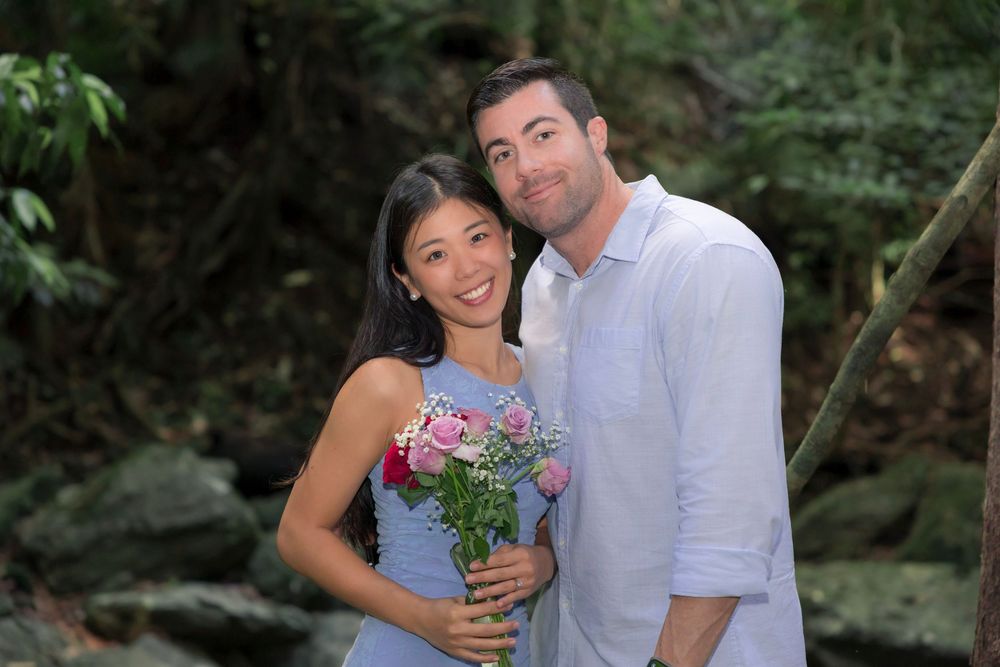 A couple poses for romantic outdoor engagement photos in a lush green forest setting with floral bouquet.