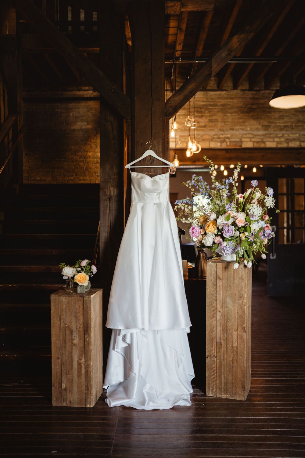 A white wedding dress hangs elegantly against a rustic wooden backdrop with floral arrangements.