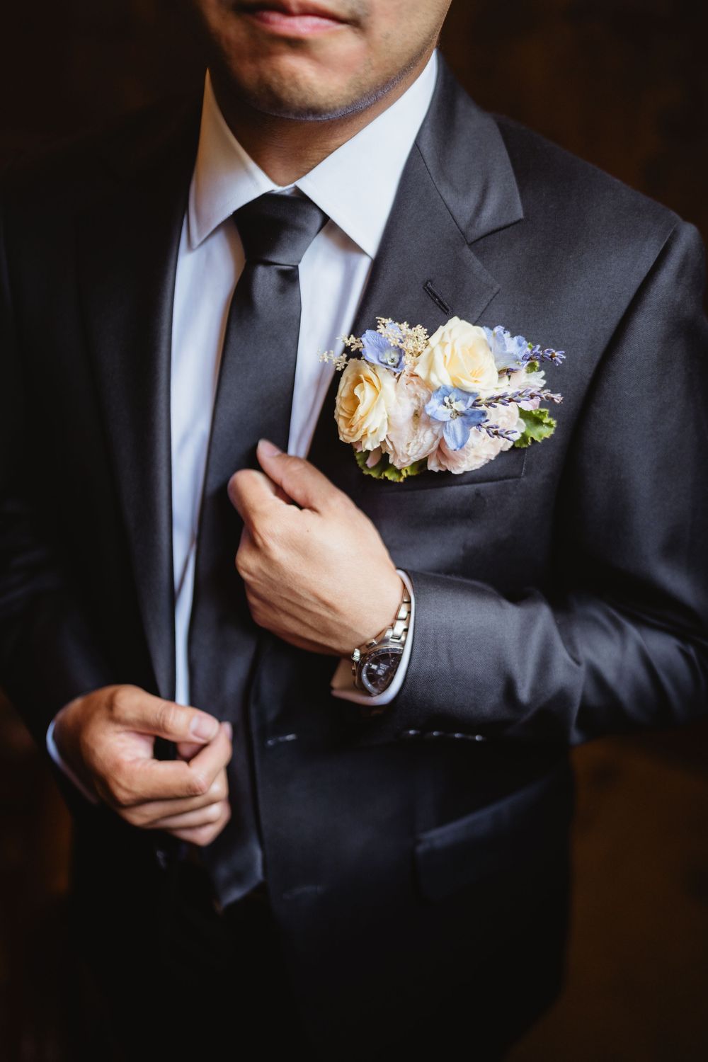 Close-up shot of wedding boutonniere with peach roses and blue flowers on a charcoal suit lapel.