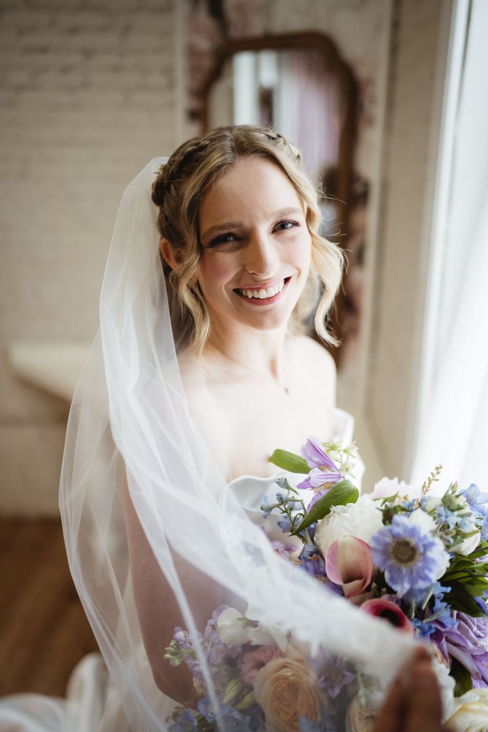 A bride in a white veil smiles while holding a colorful bouquet of pink, blue and white flowers near a window.