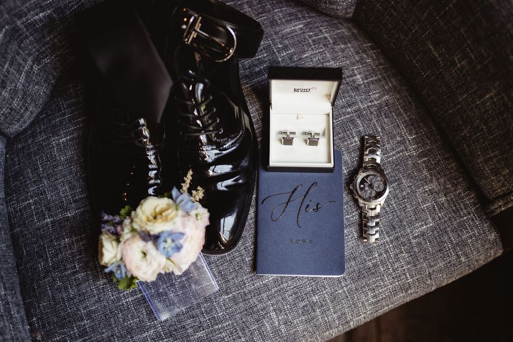 Wedding accessories including black dress shoes, diamond earrings, a navy box labeled 'His', and a white floral bouquet.