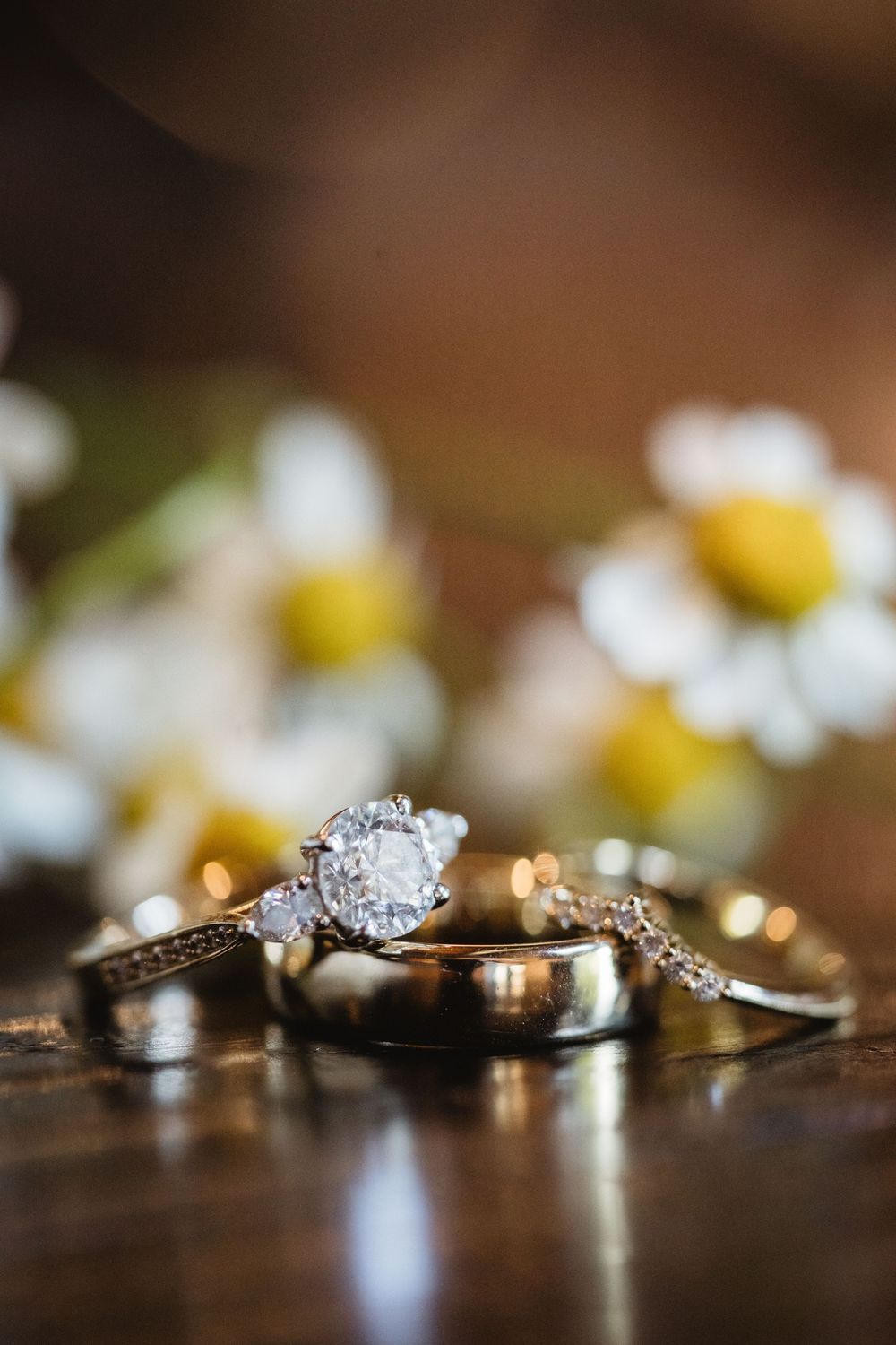 A macro shot of elegant wedding rings and diamond engagement ring displayed with soft bokeh flowers in the background.