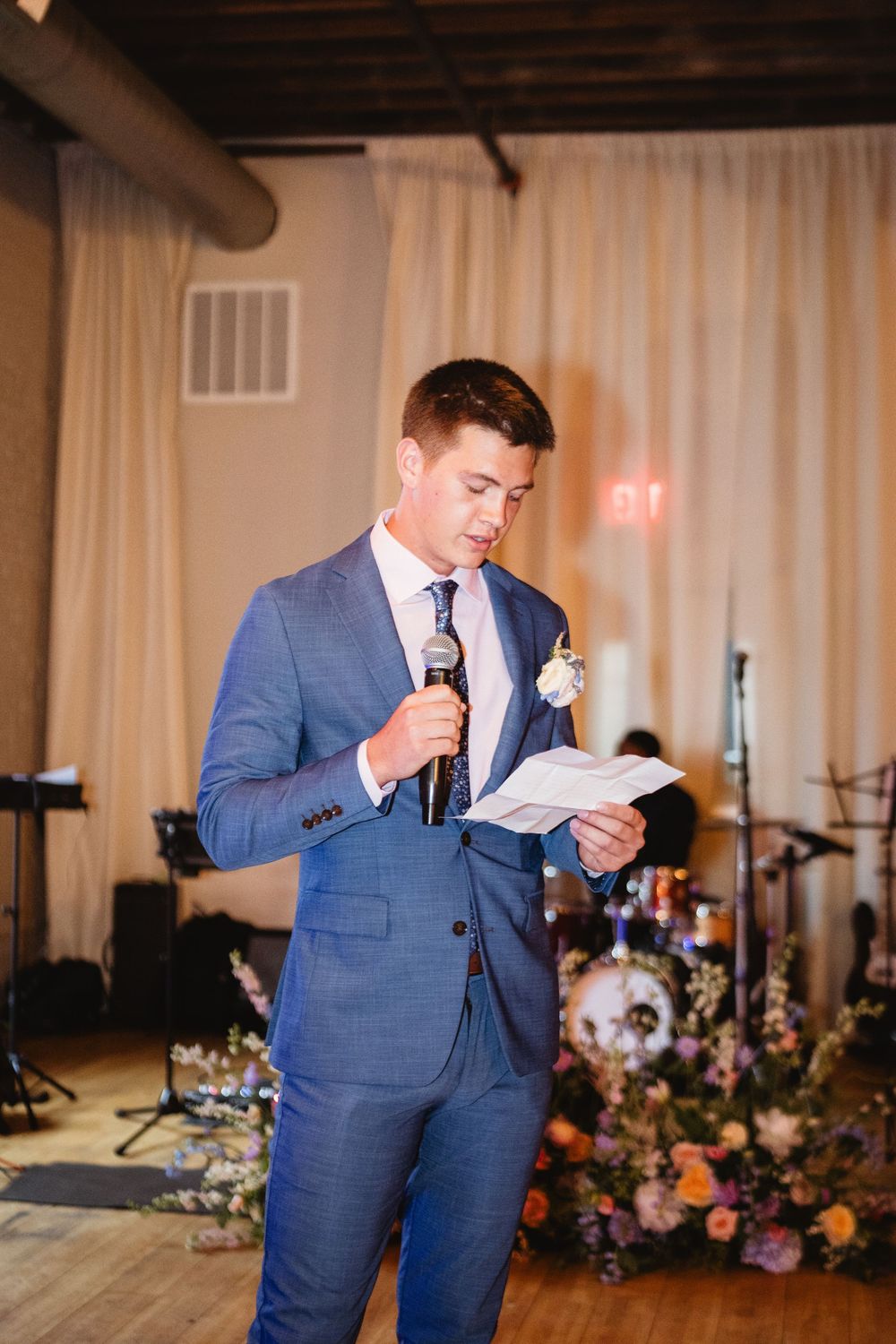 Someone in a blue suit gives a speech at a wedding reception with a floral arrangement and band setup visible in background.