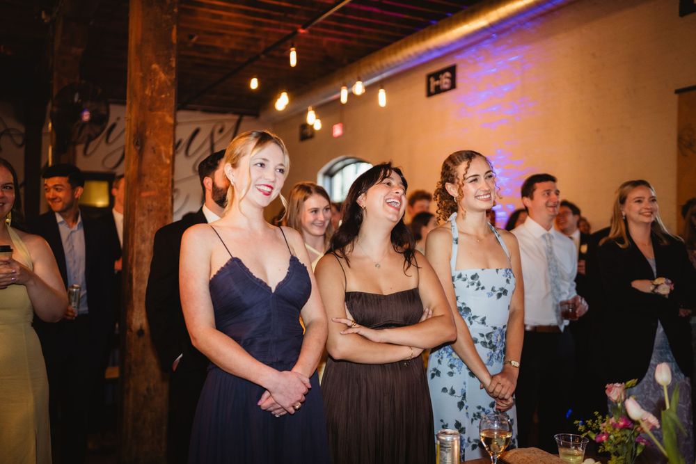 Party guests in formal attire laugh and smile together at an indoor event with string lights and exposed wooden beams.
