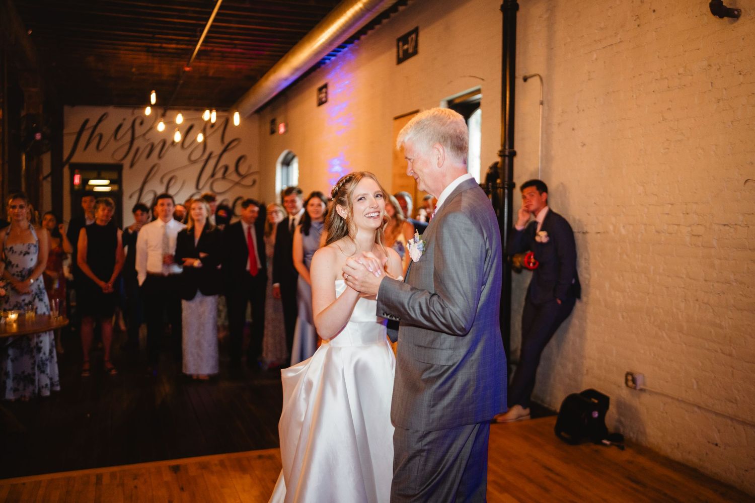 Guests dance and celebrate on a dimly lit dance floor at a wedding reception in an atmospheric venue with string lights.