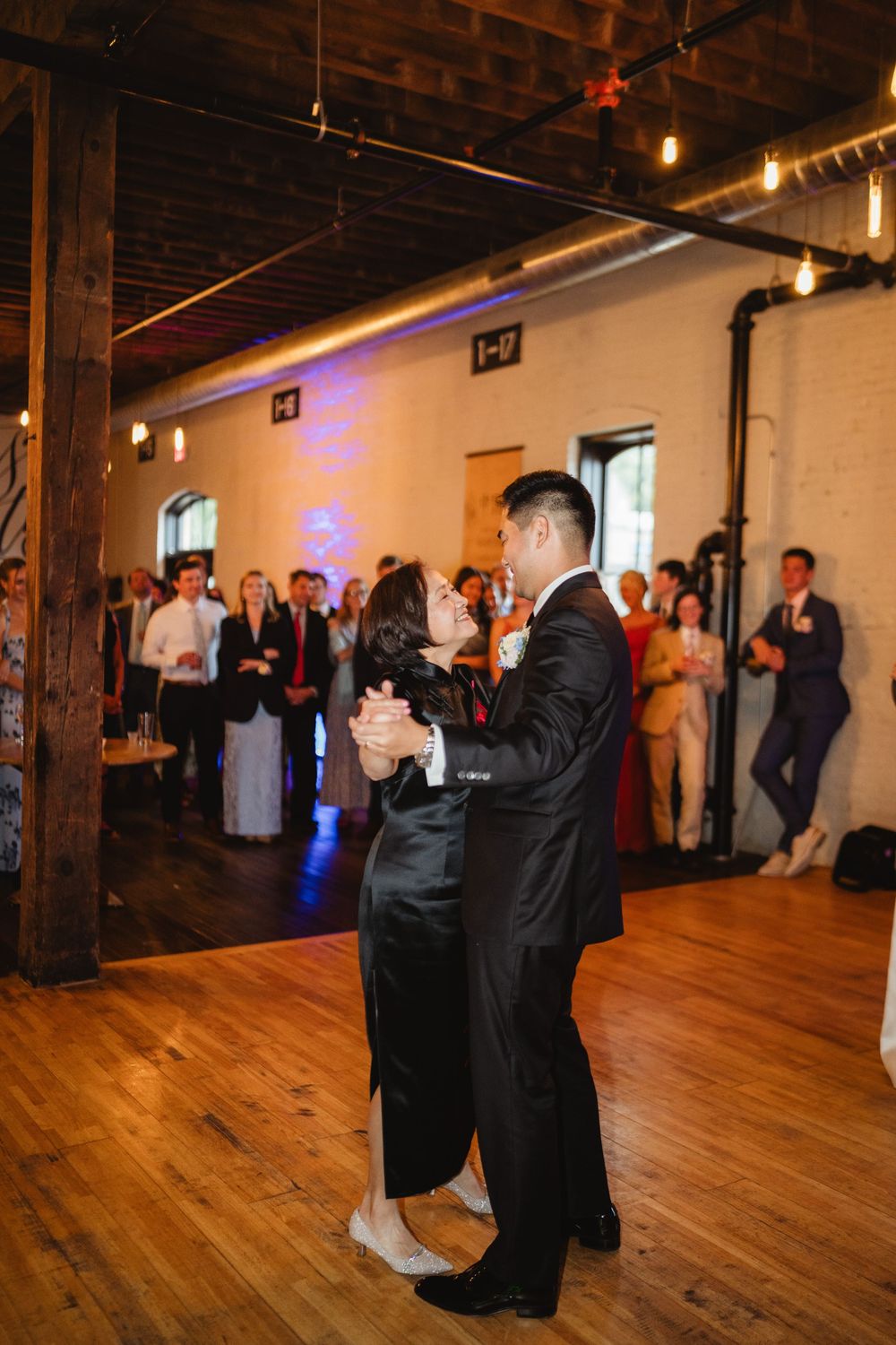 Wedding guests dance together in a spacious industrial loft venue with exposed beams and wooden floors.