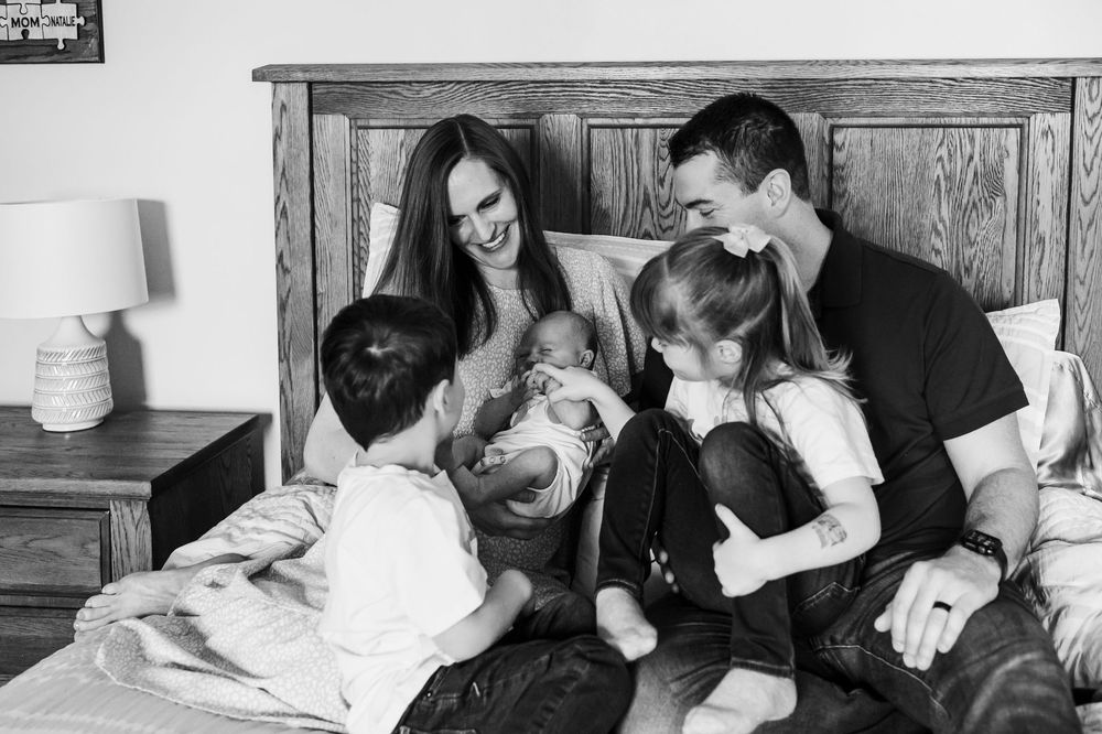 A candid black and white photograph of a family enjoying a playful moment together on a bed.