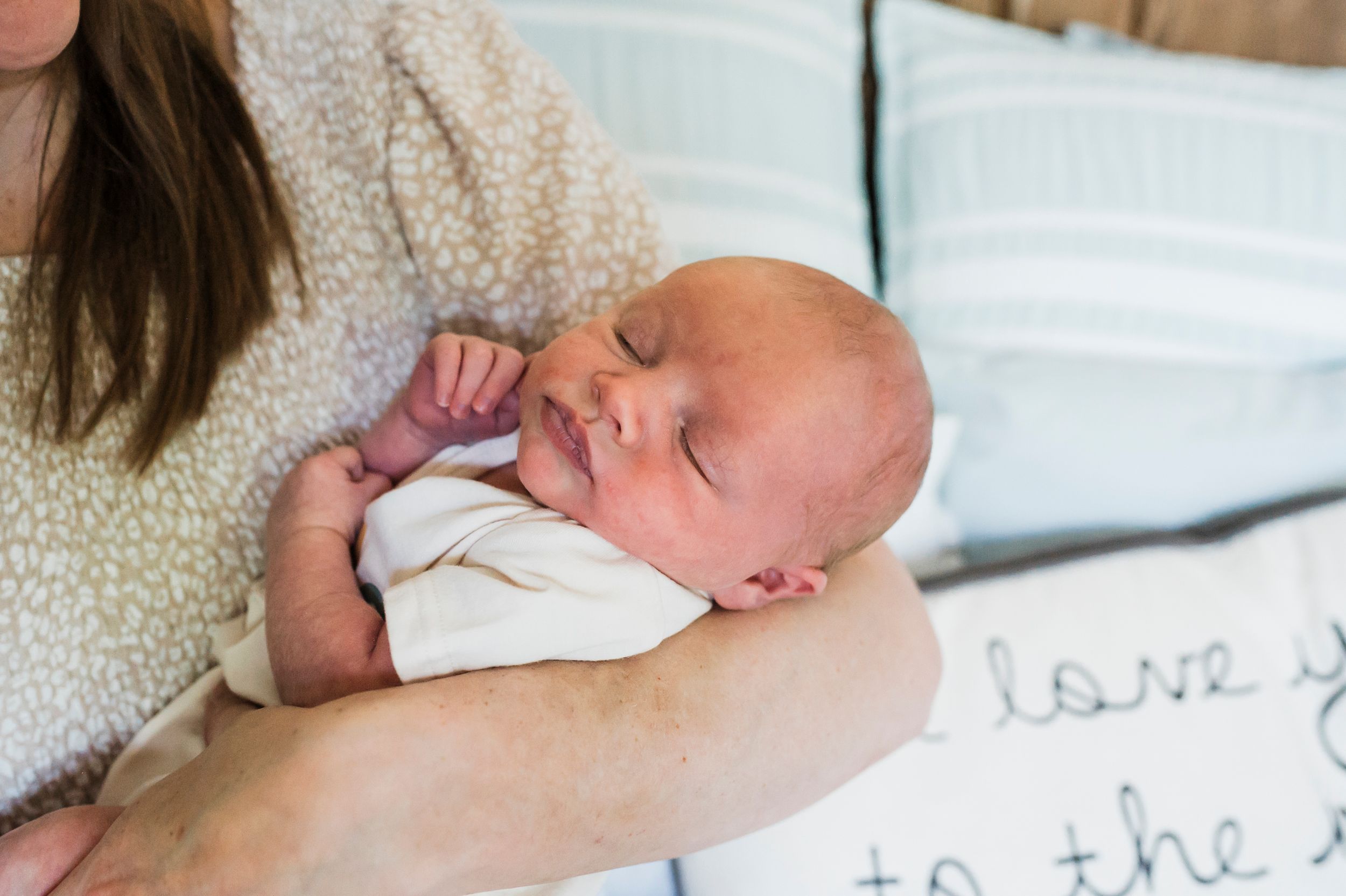 A peaceful sleeping newborn baby in a white onesie rests on someone's arm.