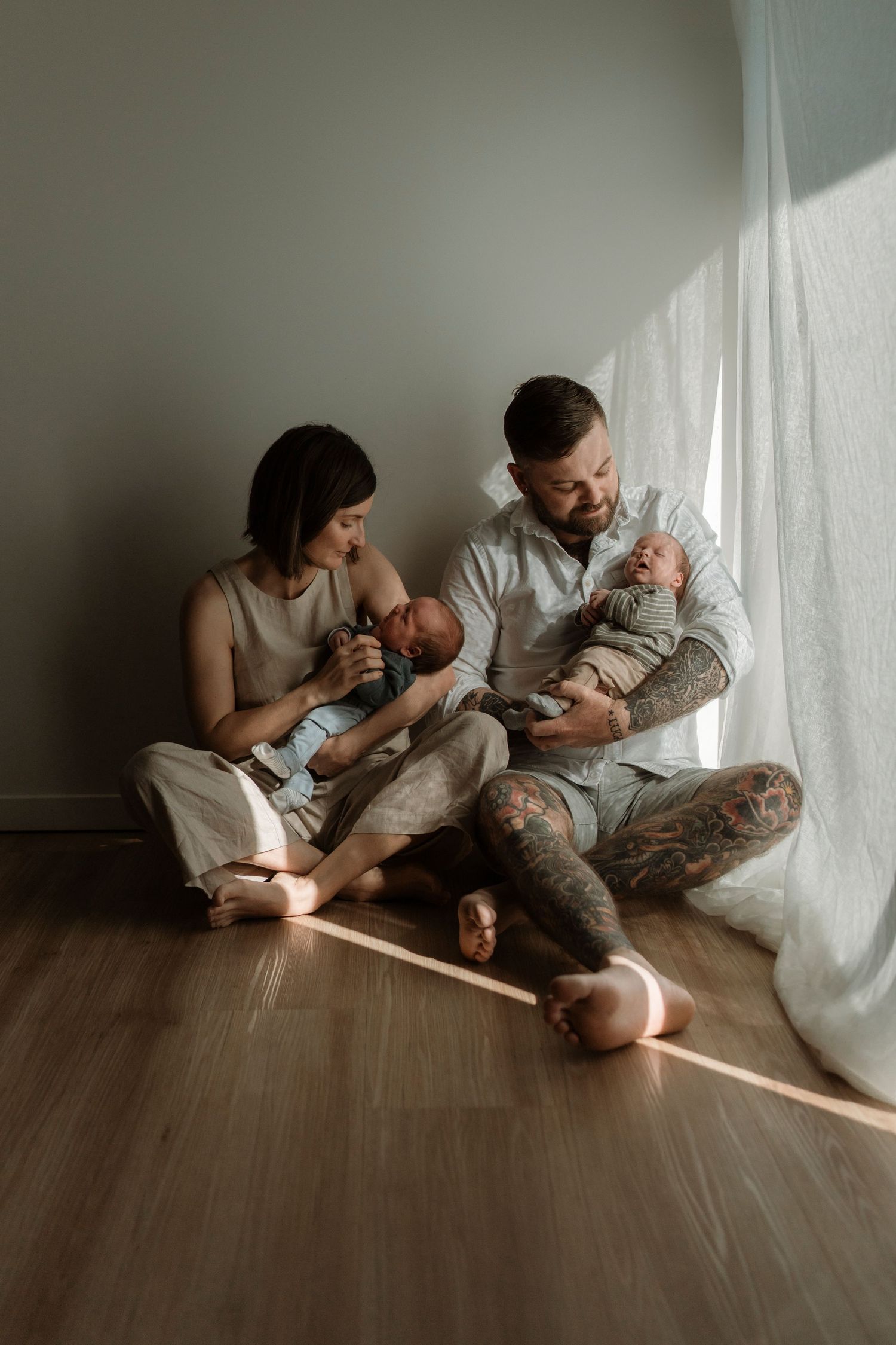 Parents sit together on hardwood floor with infant twins in a minimalist setting with natural window light.