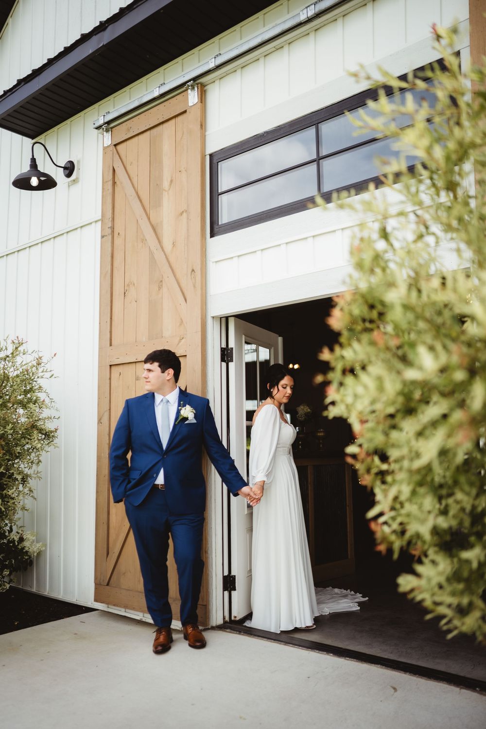 A couple in wedding attire stands on opposite sides of a barn door at a rustic venue with modern farmhouse details.