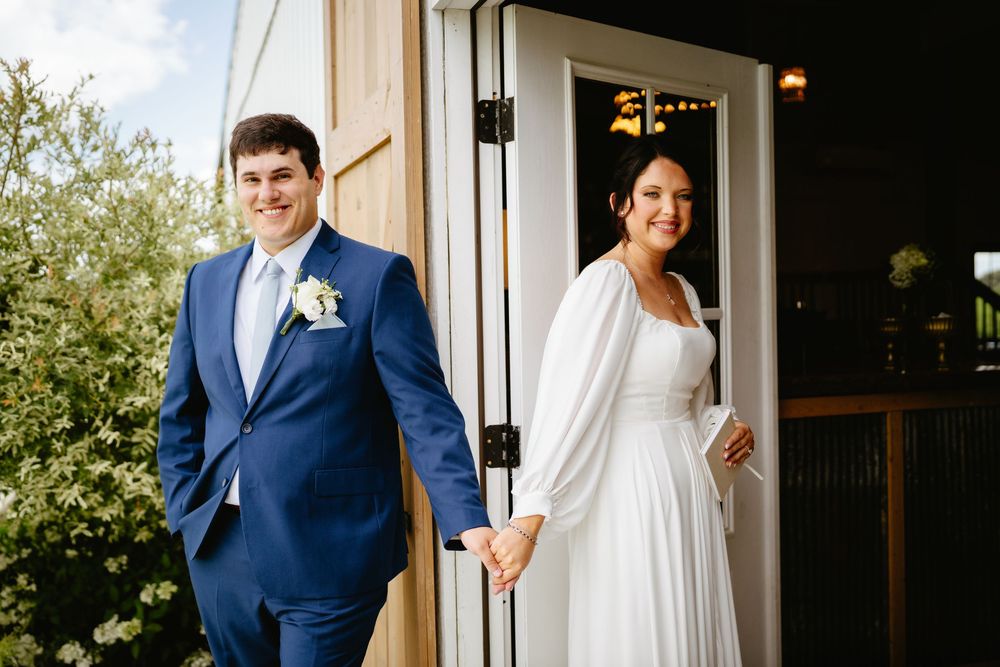 A bride and groom step out from a doorway in formal wedding attire - she in white, he in a navy blue suit.