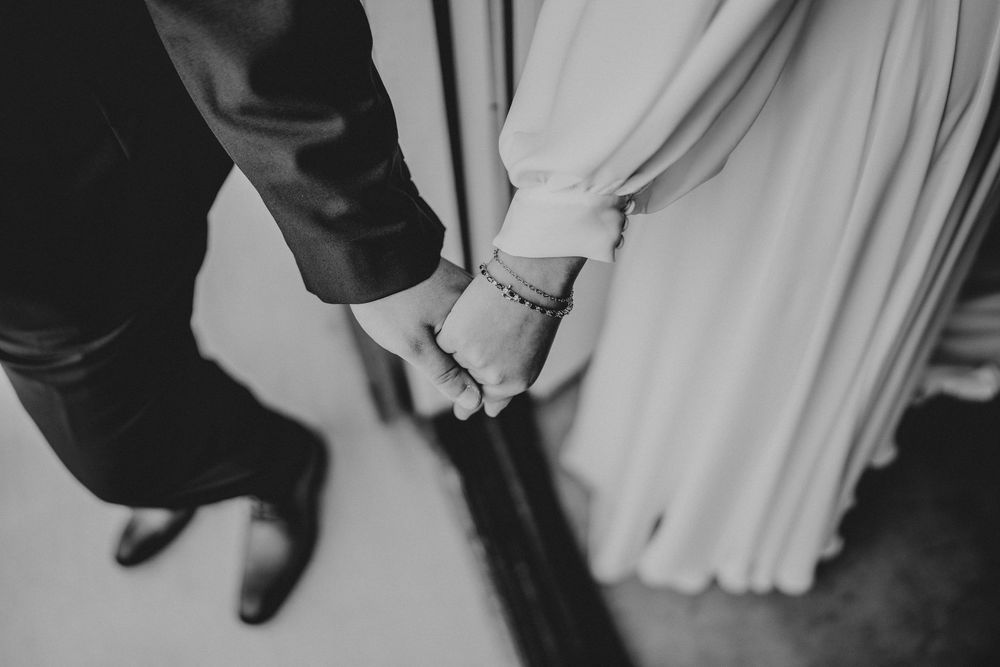 A black and white close-up photograph of a couple holding hands, showing a bride in white and groom in dark attire.
