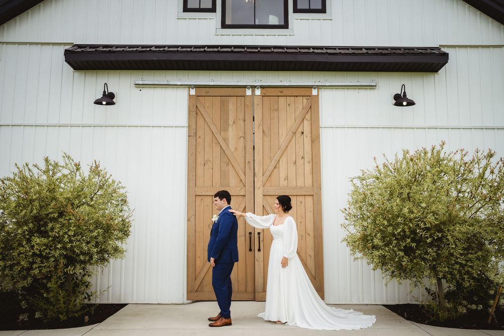 Two people in formal attire stand facing a rustic barn door with decorative shrubs on either side.