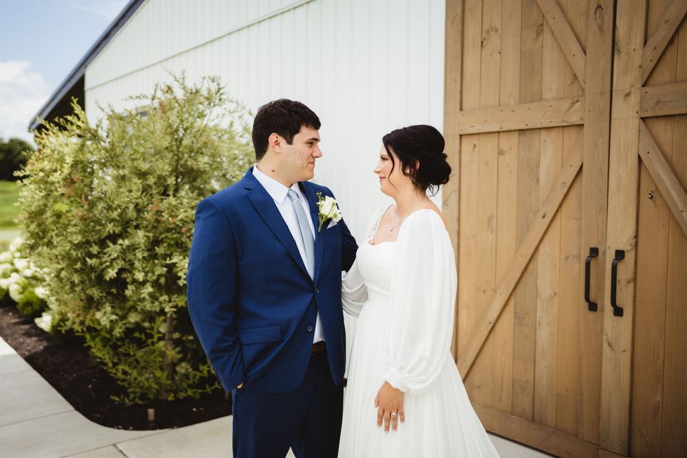 A wedding couple stands together in front of a rustic barn door wearing traditional formal wedding attire.